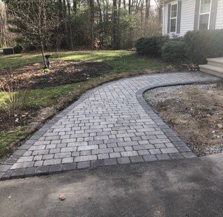 Curved gray paver walkway beside a house, leading from a driveway into a wooded yard.