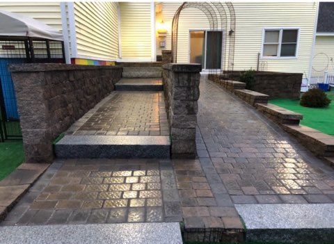 Stone patio and walkway with brick steps leading to a house entrance, bordered by low walls and planters.