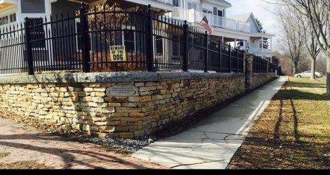 Stone retaining wall and black iron fence along a sidewalk beside a house in a sunny residential yard