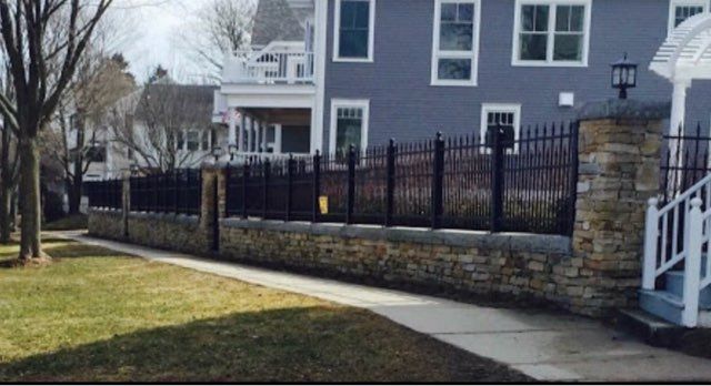 Residential house with black iron fence, stone wall, and sidewalk beside a grassy yard