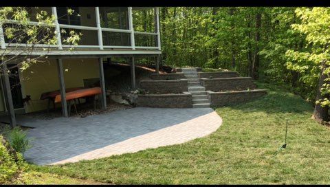 Backyard patio with stone steps, raised deck, and grassy yard beside wooded trees