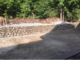Stone retaining wall beside a dirt road, with trees and sunlight casting shadows.