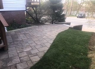 Stone patio beside a green lawn and front steps, with shrubs and a driveway in the background
