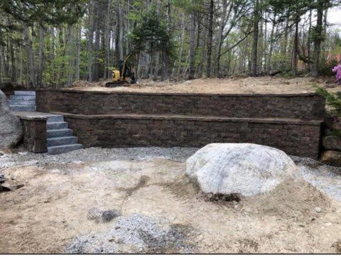 Terraced stone retaining wall in a wooded yard with steps, boulders, and a small excavator above the wall