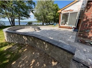 Curved stone patio beside a lake, with trees, brick house, and glass door.