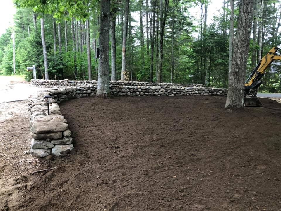 Freshly graded soil in a wooded yard beside a stone retaining wall, with an excavator at right