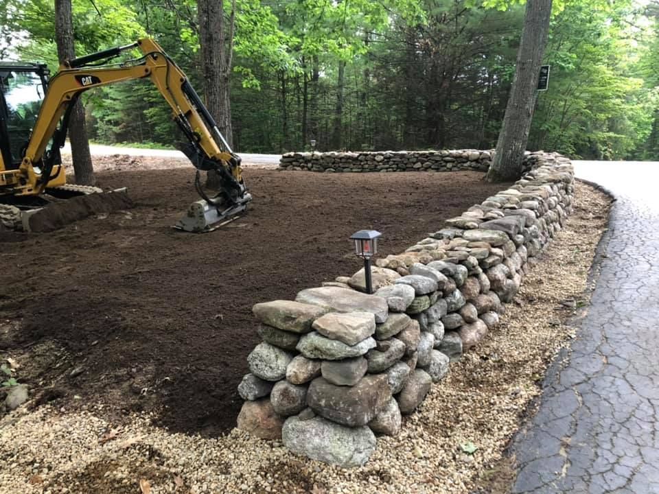 Yellow excavator working beside a stone retaining wall on a wooded roadside.
