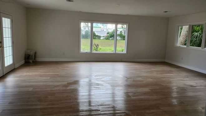 Empty living room with hardwood floors, large windows, and a view of greenery outside.