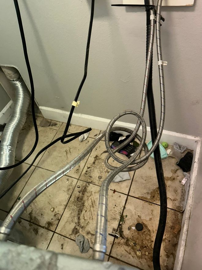 Cluttered utility area with exposed pipes, cables, and water-stained flooring near a wall.
