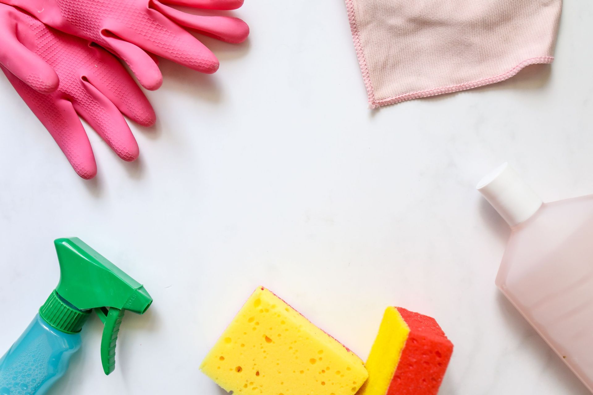Cleaning supplies on a white surface, including pink gloves, spray bottle, sponge, and cloth.