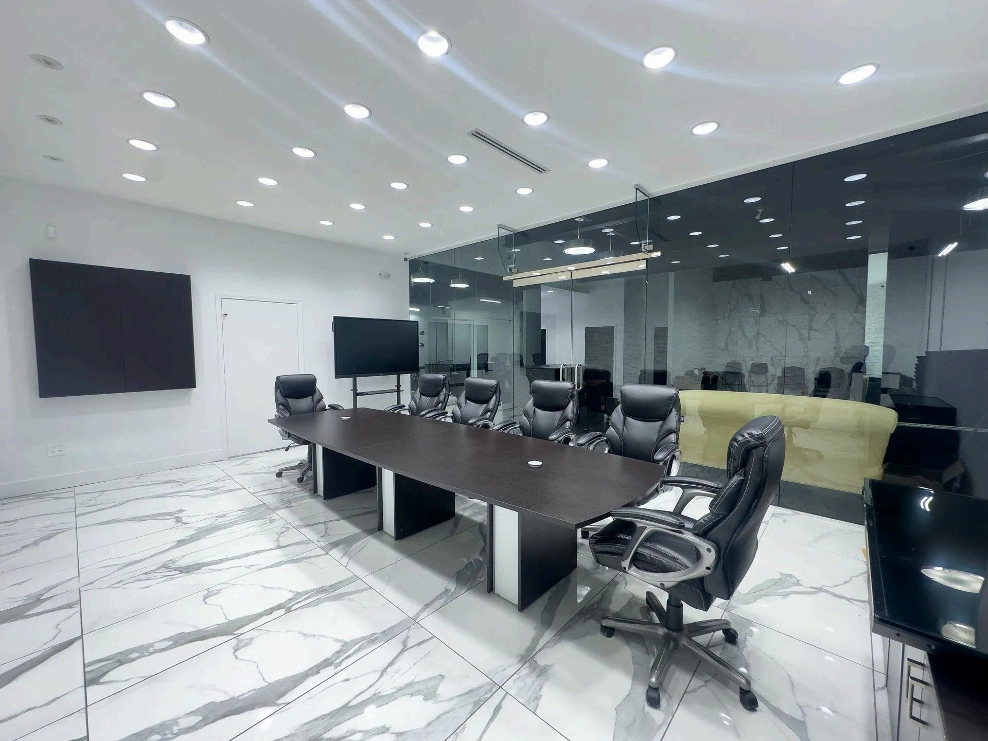 Modern conference room with long table, black chairs, wall screens, and glass walls under bright ceiling lights