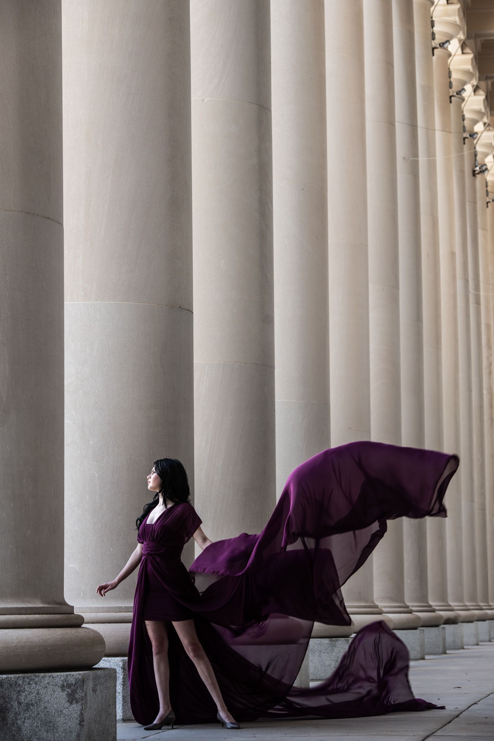Woman in a flowing purple dress walking beside tall white columns, the train billowing behind her.