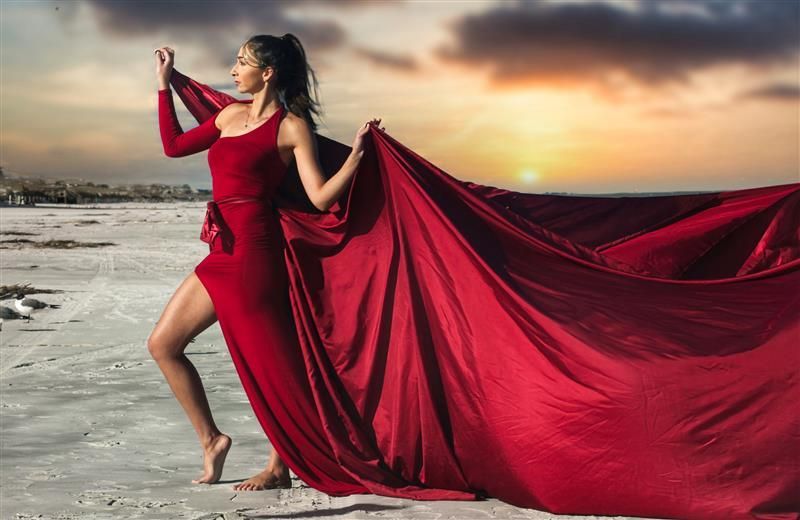 Woman in a flowing red dress posing on a beach at sunset, with fabric billowing in the wind.