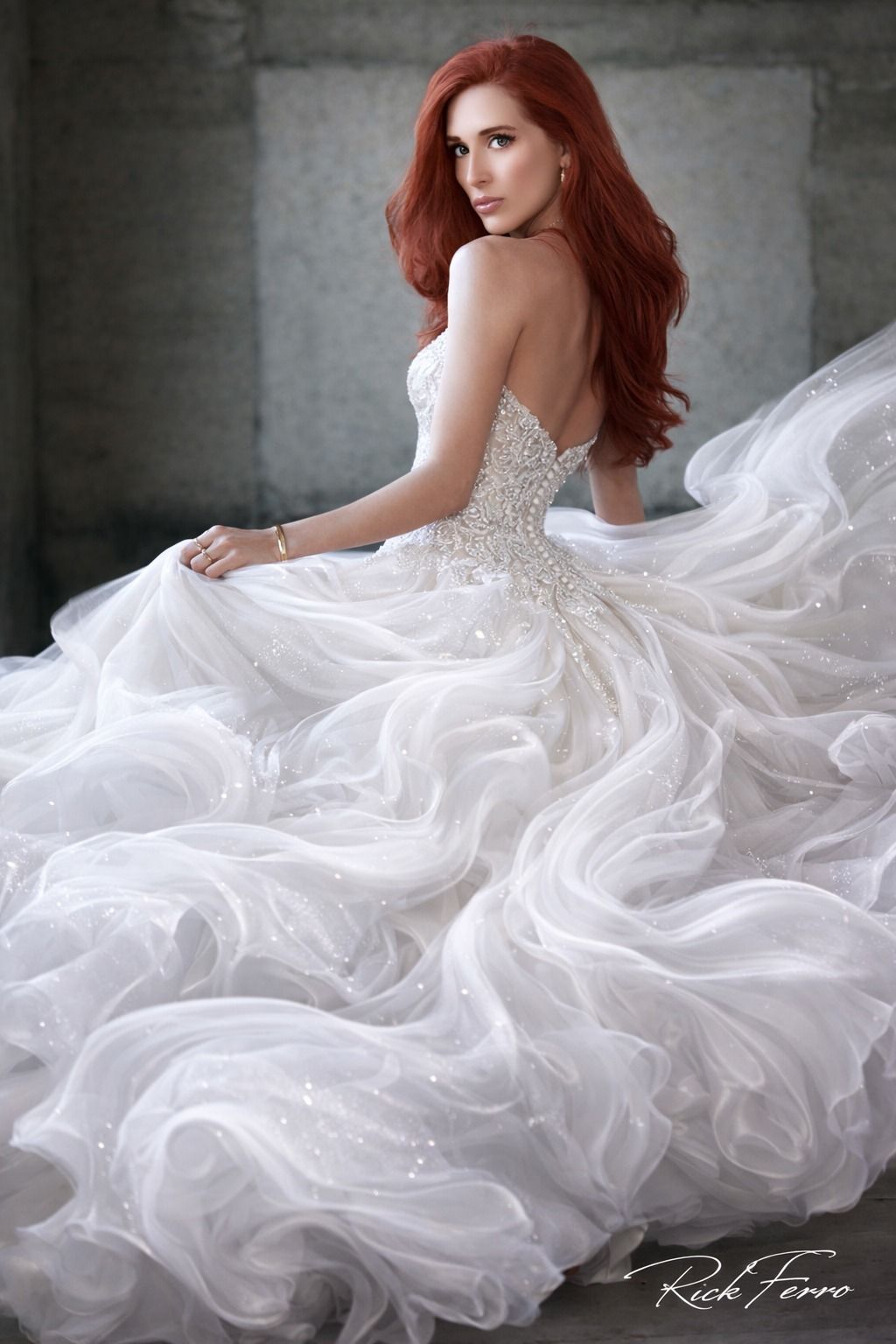 Woman in a white ruffled gown seated against a gray backdrop, looking over her shoulder.