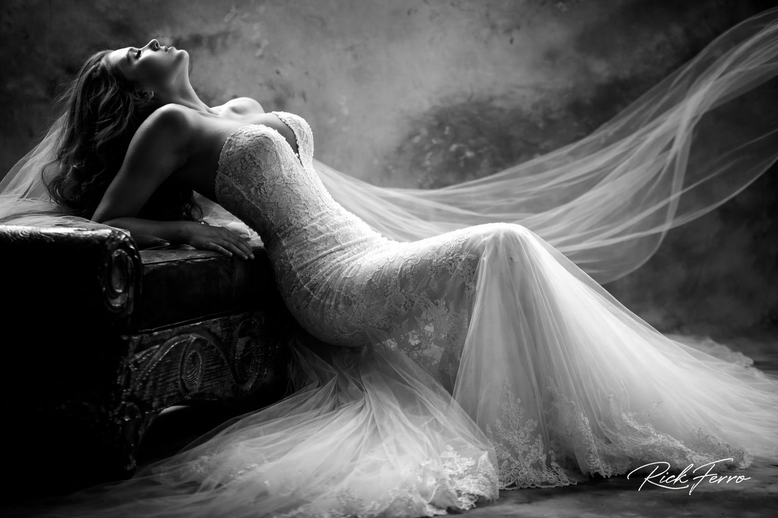 Woman in a white gown reclining on a couch, draped in flowing sheer fabric, in a dramatic black-and-white scene