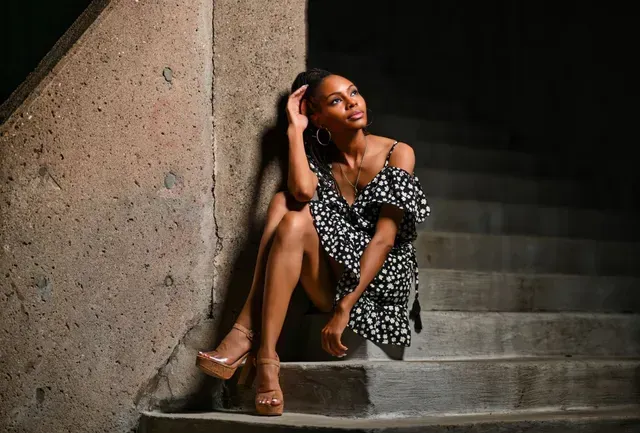 Woman in a black floral dress sitting on concrete steps, leaning against a wall and looking upward.