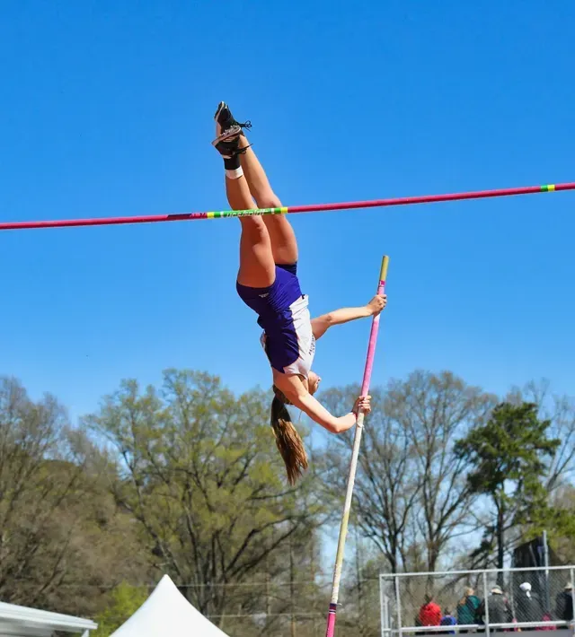 Pole vaulter upside down over the bar outdoors, in a purple uniform with blue sky behind.