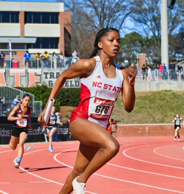 NC State runner sprints on a red track, baton in hand, during a relay race.