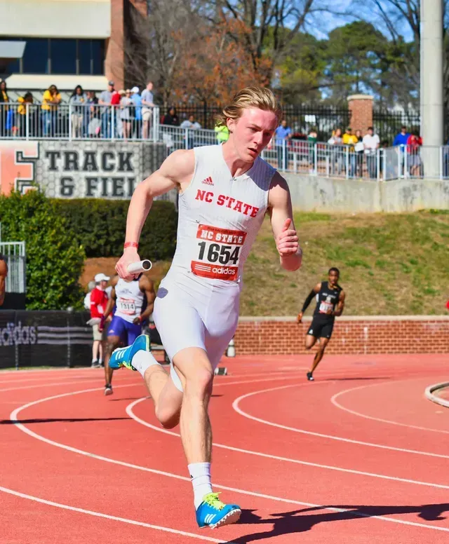 Runner in white NC State uniform leading a track race on a sunny day