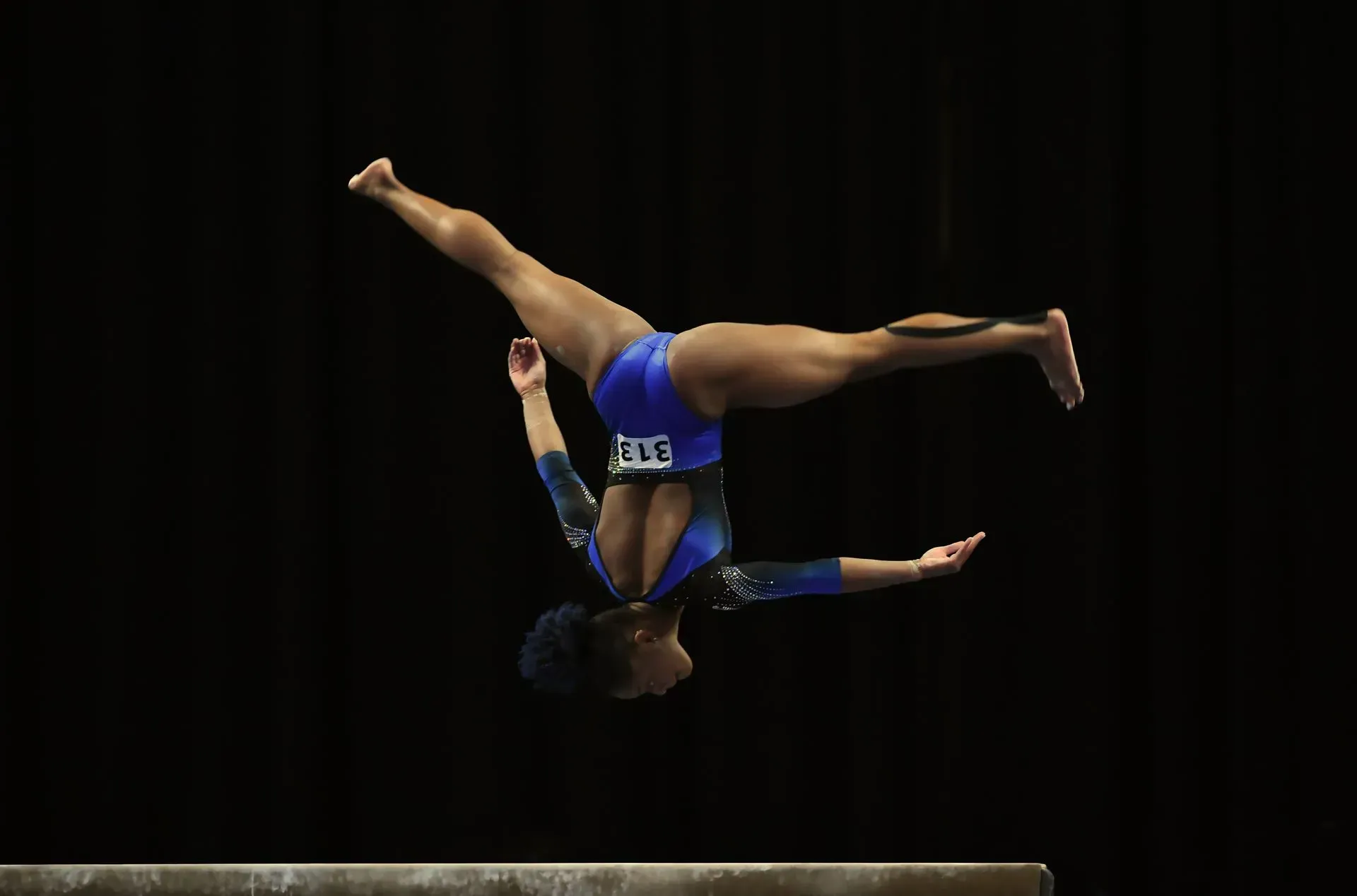 Gymnast in blue leotard performing a handstand split on a balance beam against a black background