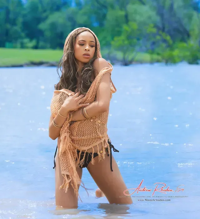 Woman standing in shallow blue water, wearing a tan knit cover-up against a green shoreline.