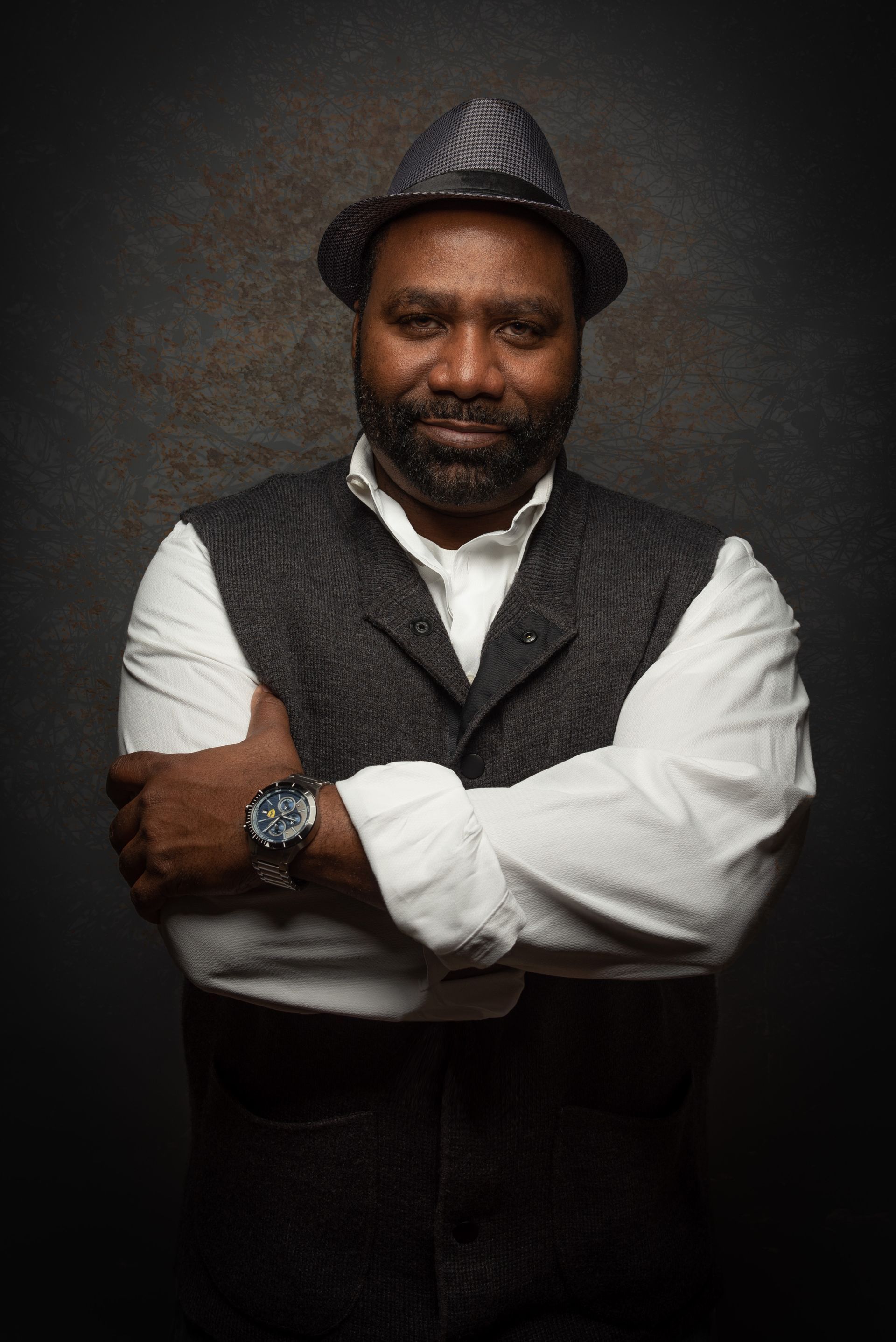 Portrait of a man in a gray hat and vest, arms crossed, standing against a dark studio backdrop