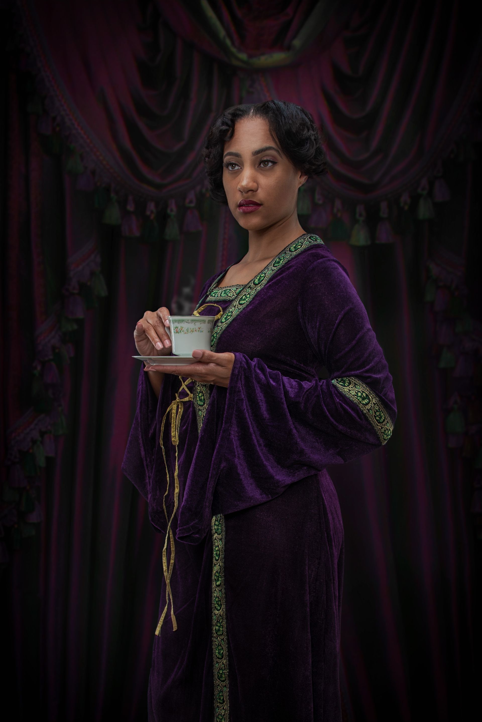 Woman in a purple robe holding a cup, standing against dark draped fabric.