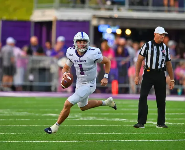 Football player in white runs with the ball on a field, with a referee nearby and blurred crowd in the background