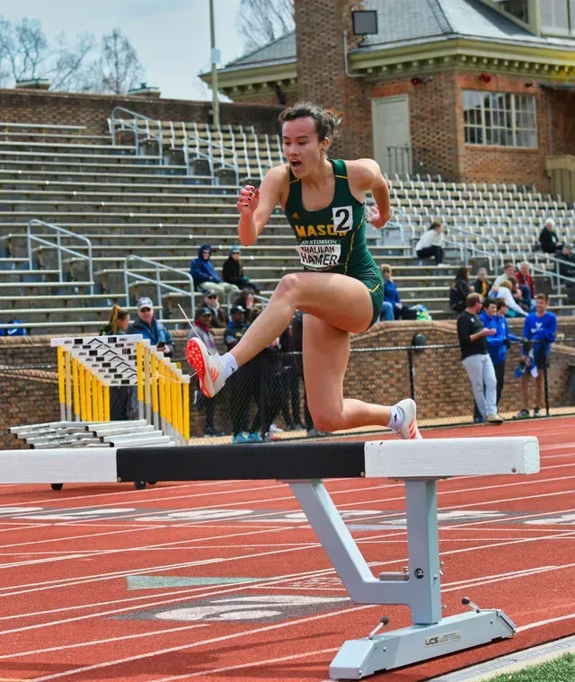 Track athlete clearing a hurdle on a stadium track, mid-jump in black uniform