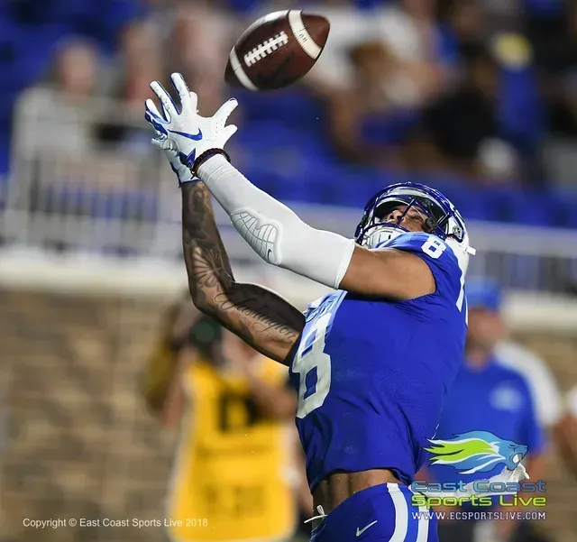 Blue-uniformed football player reaches up to catch a spiraling pass on the field