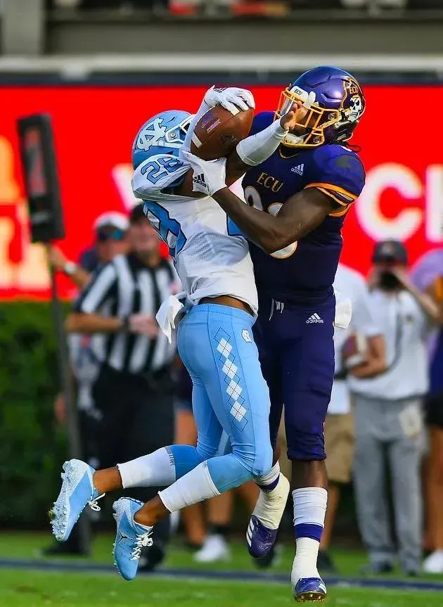 Two football players collide midair during a game, one in purple tackling one in light blue.