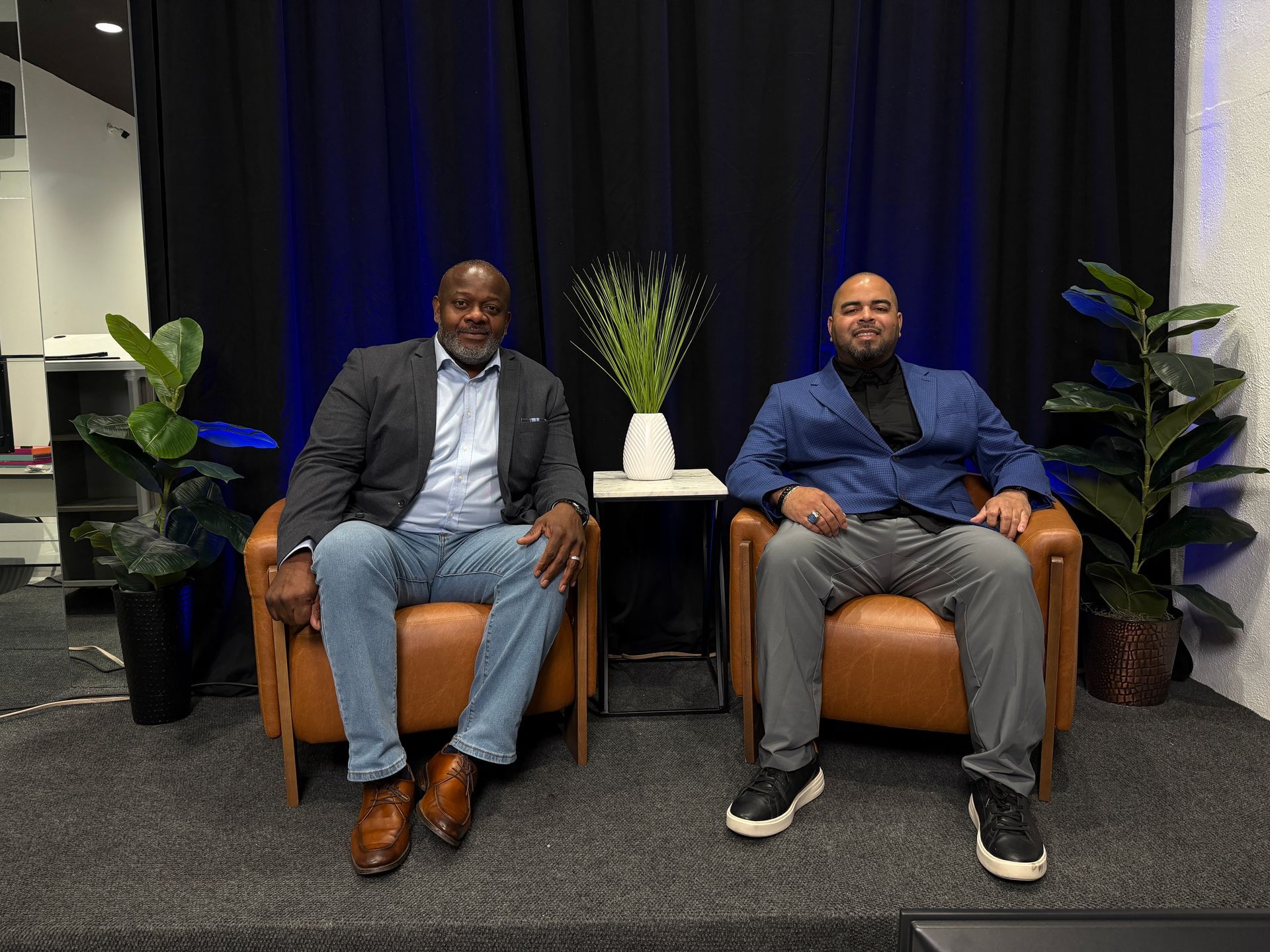 Two men seated on chairs in a lounge area with plants and a dark curtain backdrop.