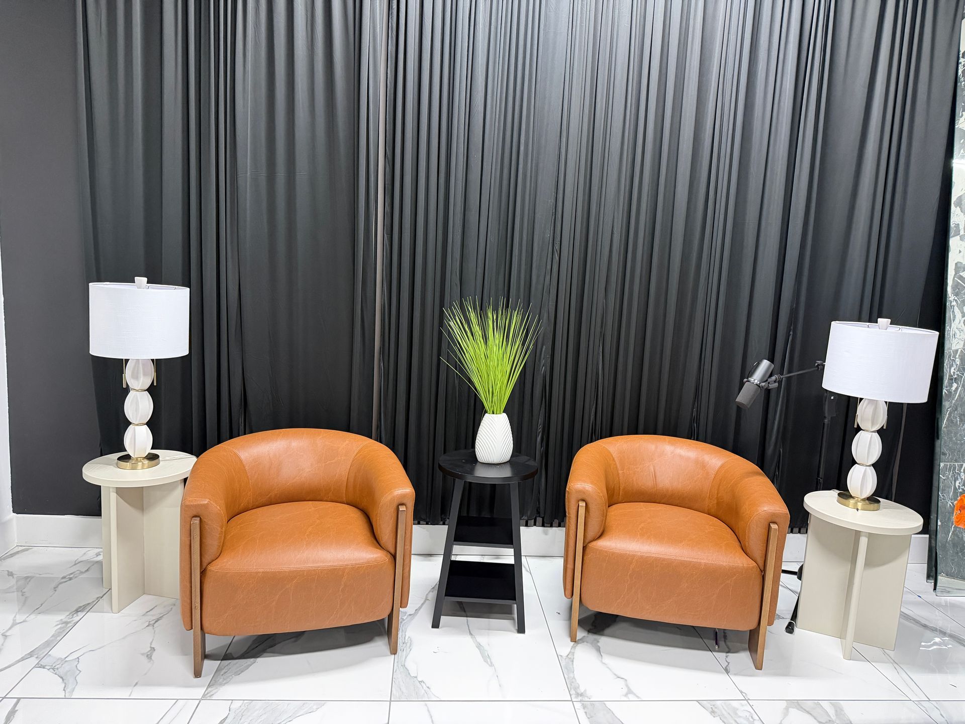 Elegant lounge with two orange chairs, black curtain backdrop, white side tables, and lamps.