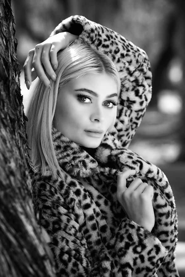 Black-and-white portrait of a person in a leopard-print coat, posing with one hand over their head.