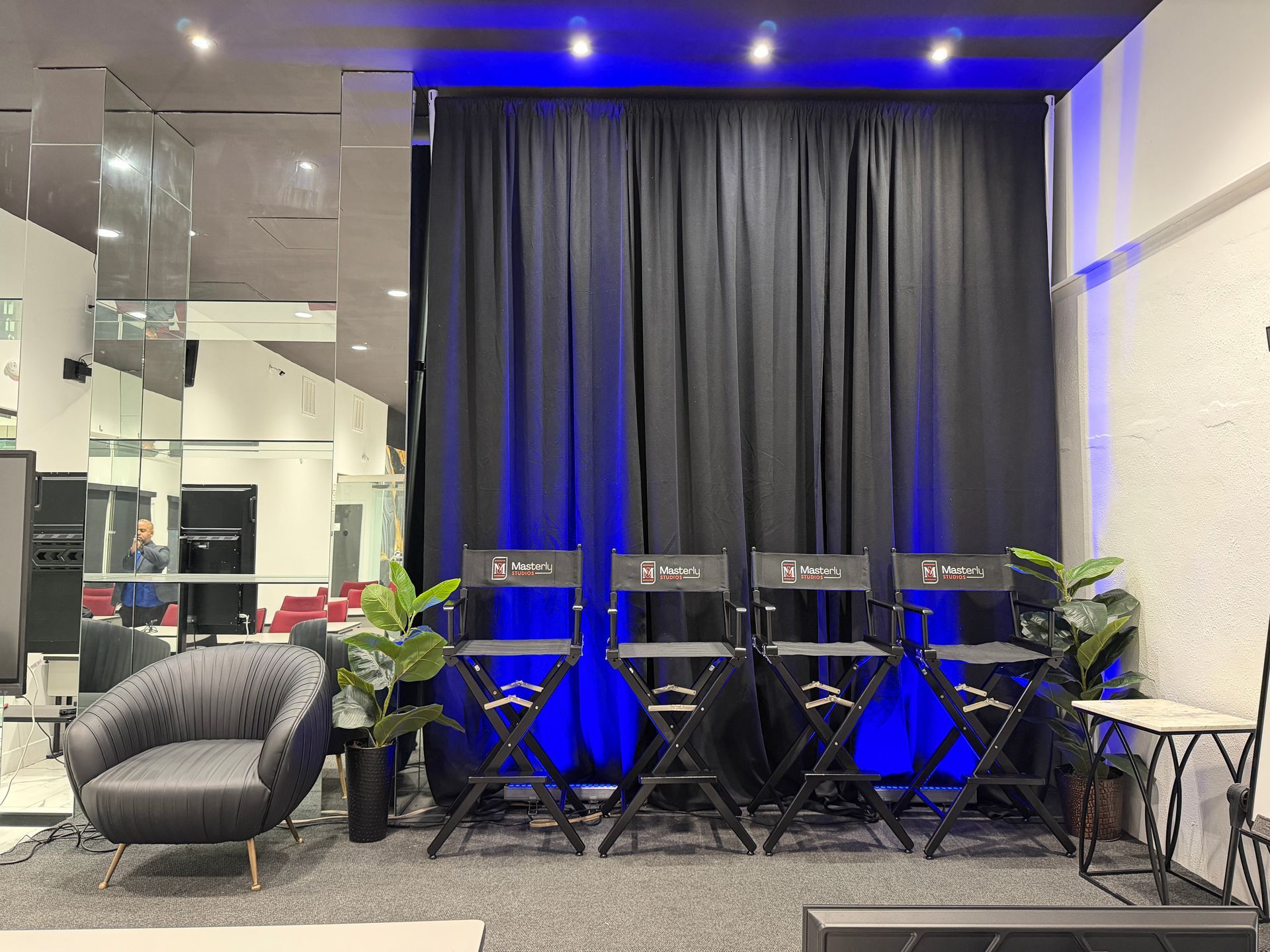 Blue-lit lounge with a curtain backdrop, folding chairs, potted plants, and a gray beanbag by mirrored walls