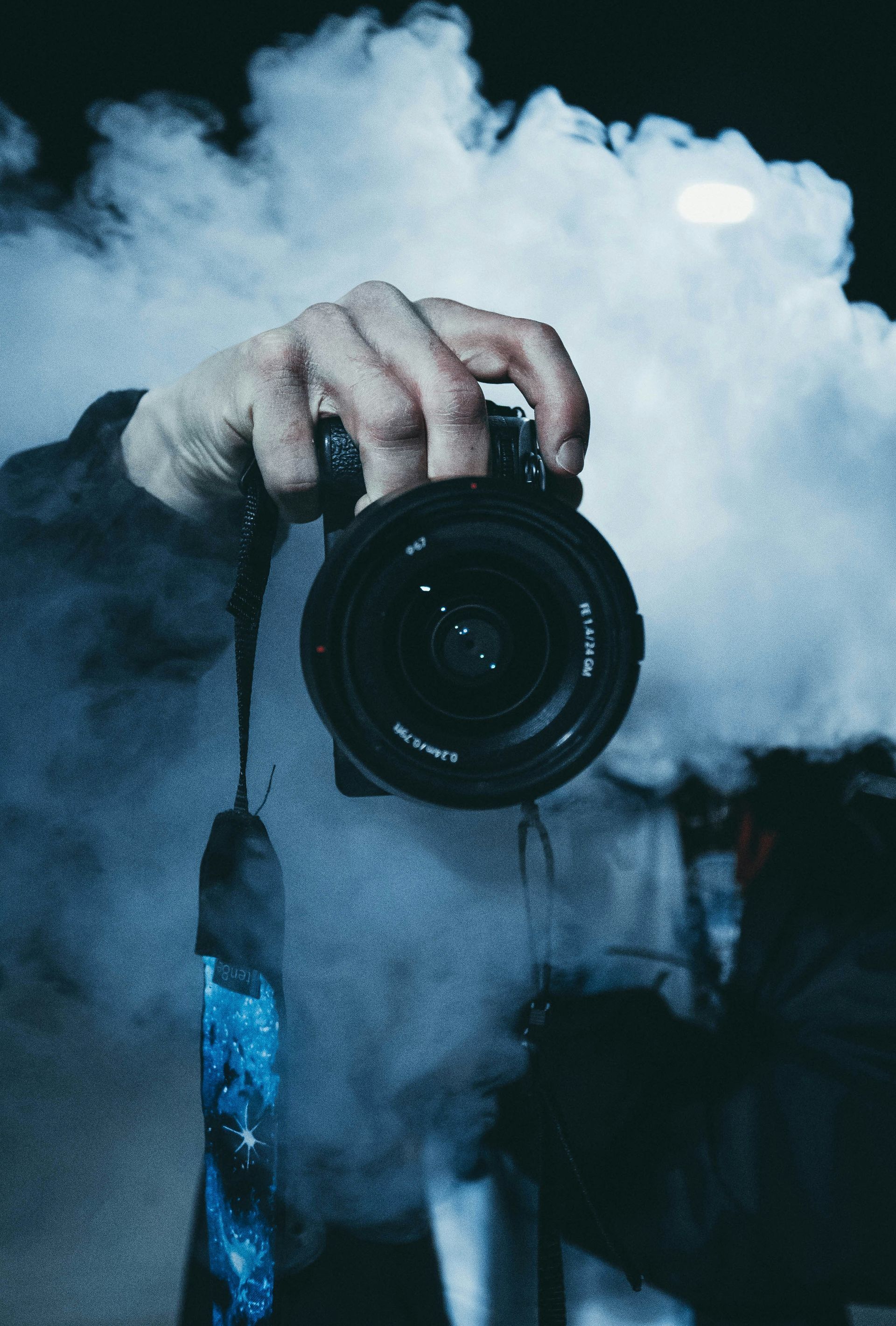 Person holding a camera amid blue smoke or fog, with the lens facing forward
