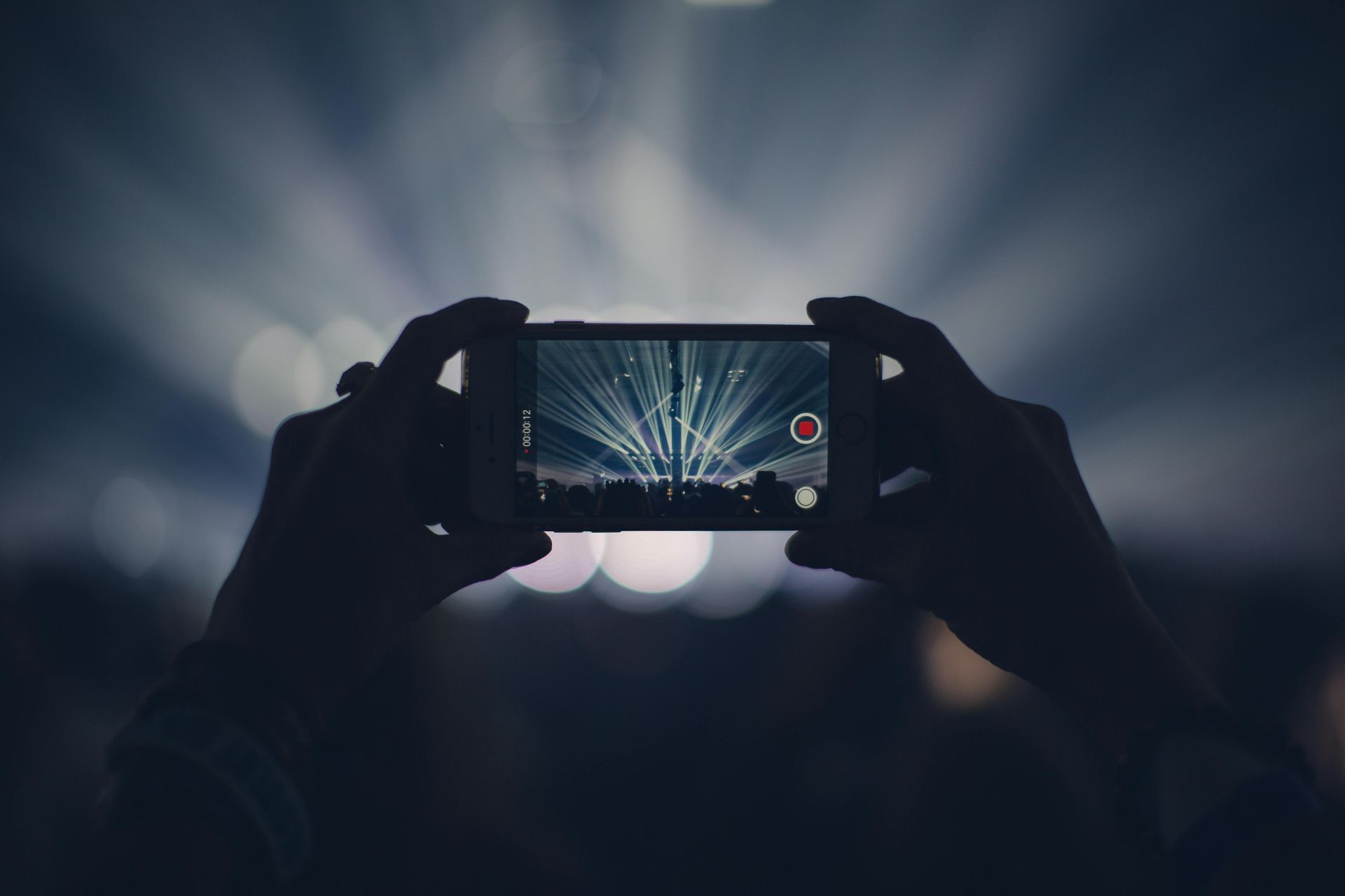 Hands holding a smartphone filming bright stage lights in a dark concert crowd.