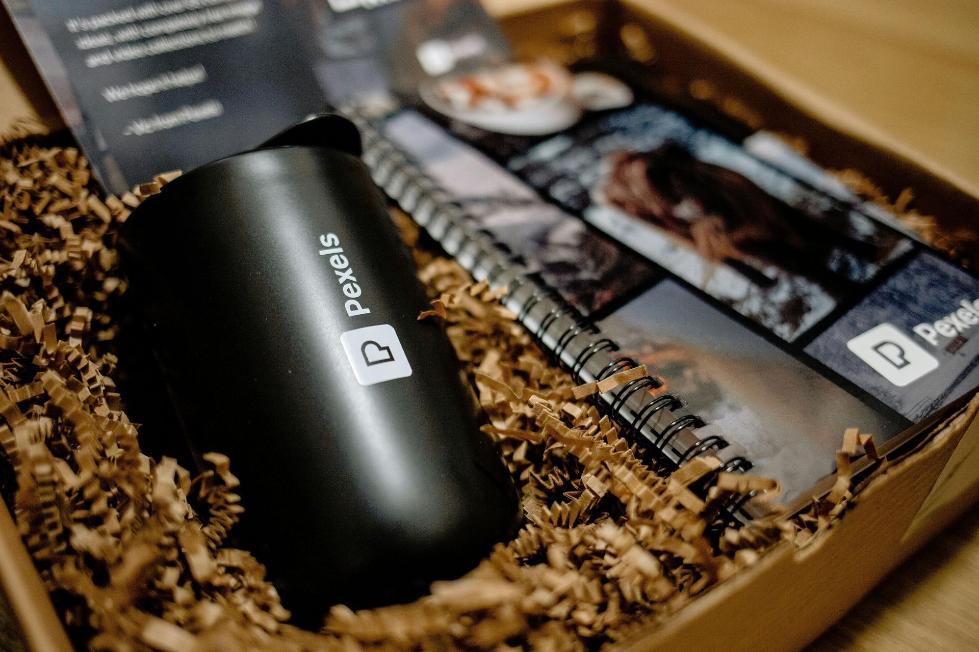 Black Pebble mouse in a straw-filled gift box beside a printed brochure and spiral-bound booklet