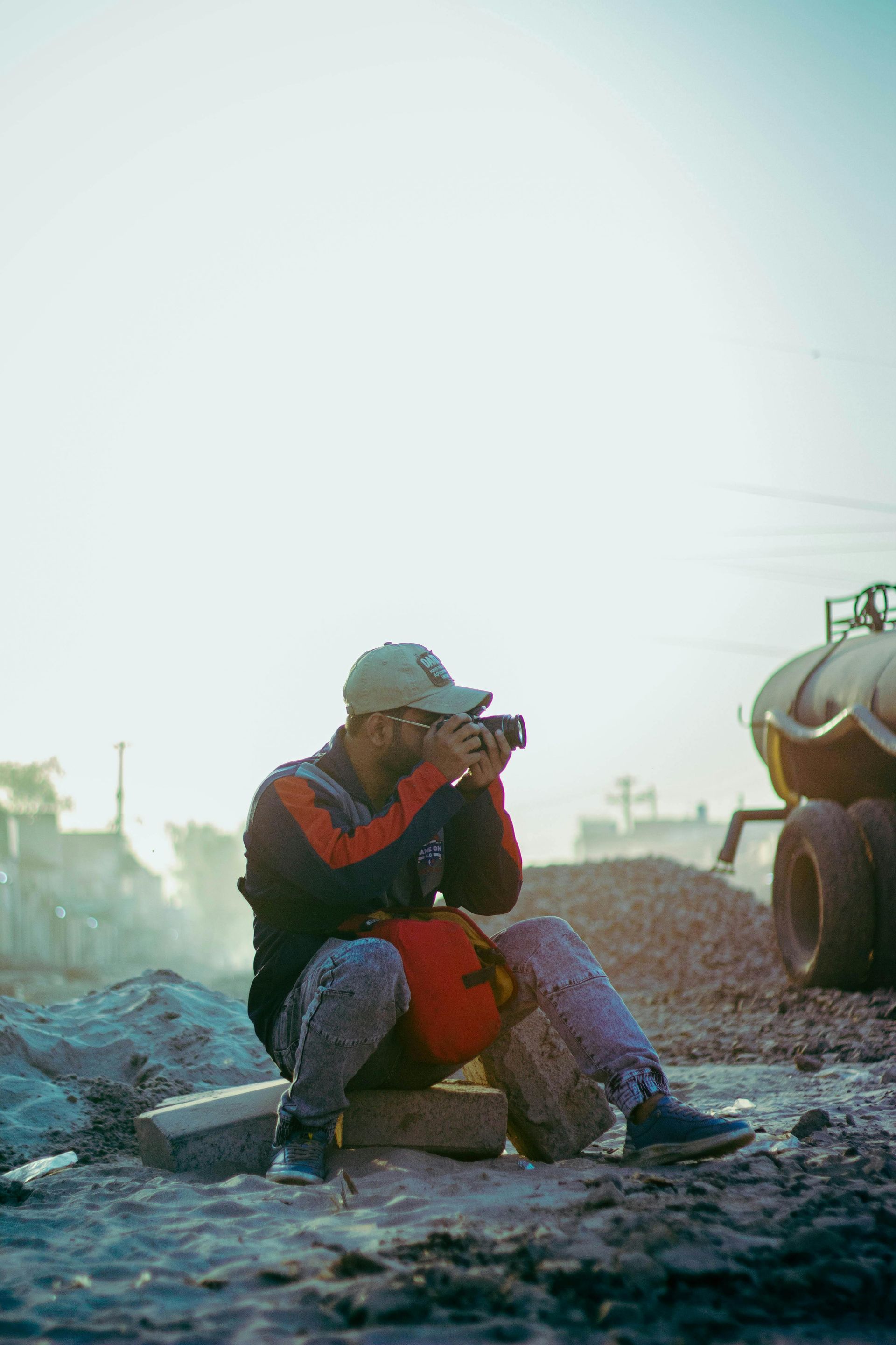 Person in a red jacket and cap sits on rocky ground, taking a photo with a camera near industrial equipment.