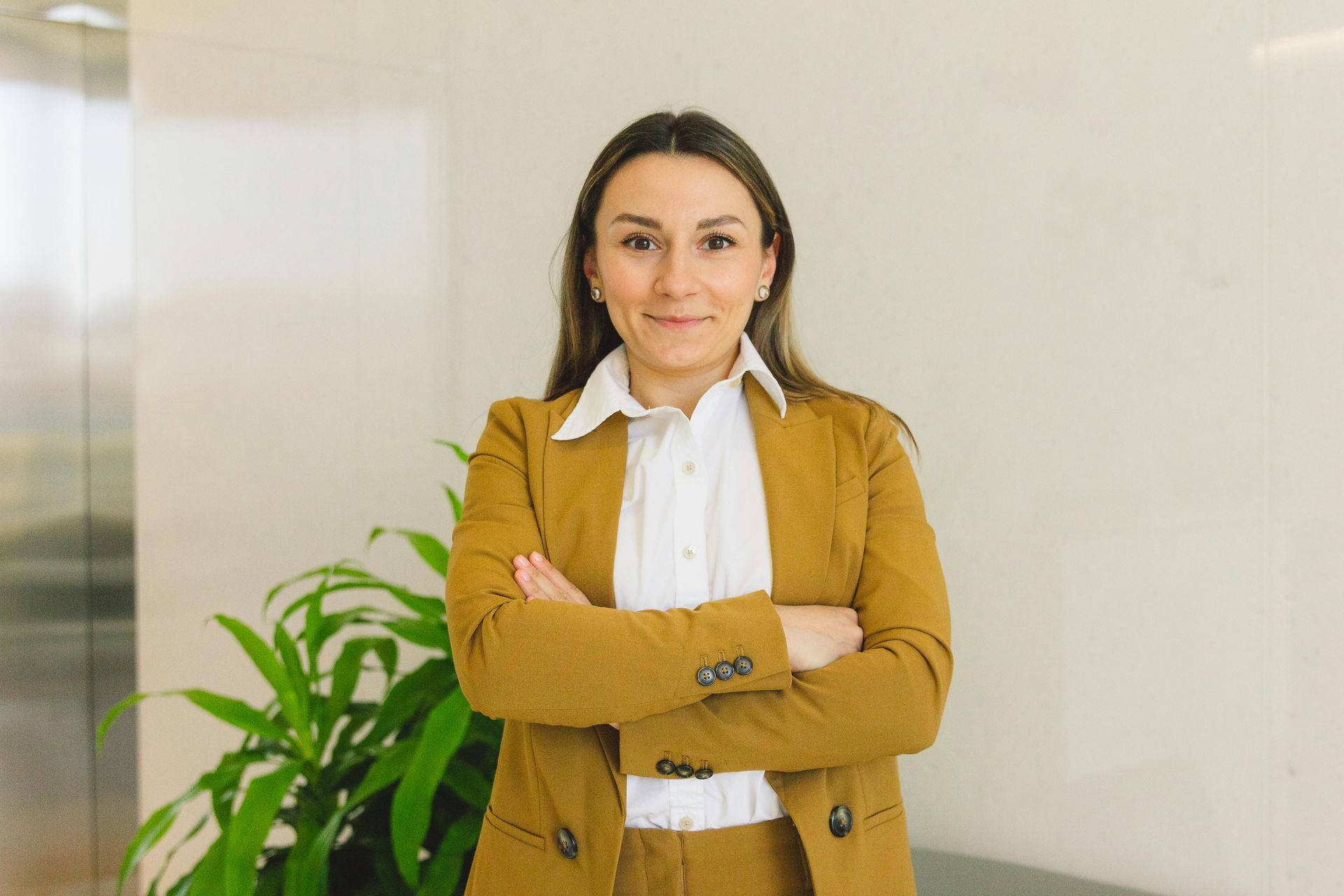 Woman with arms crossed in mustard blazer and white shirt standing indoors beside a green plant
