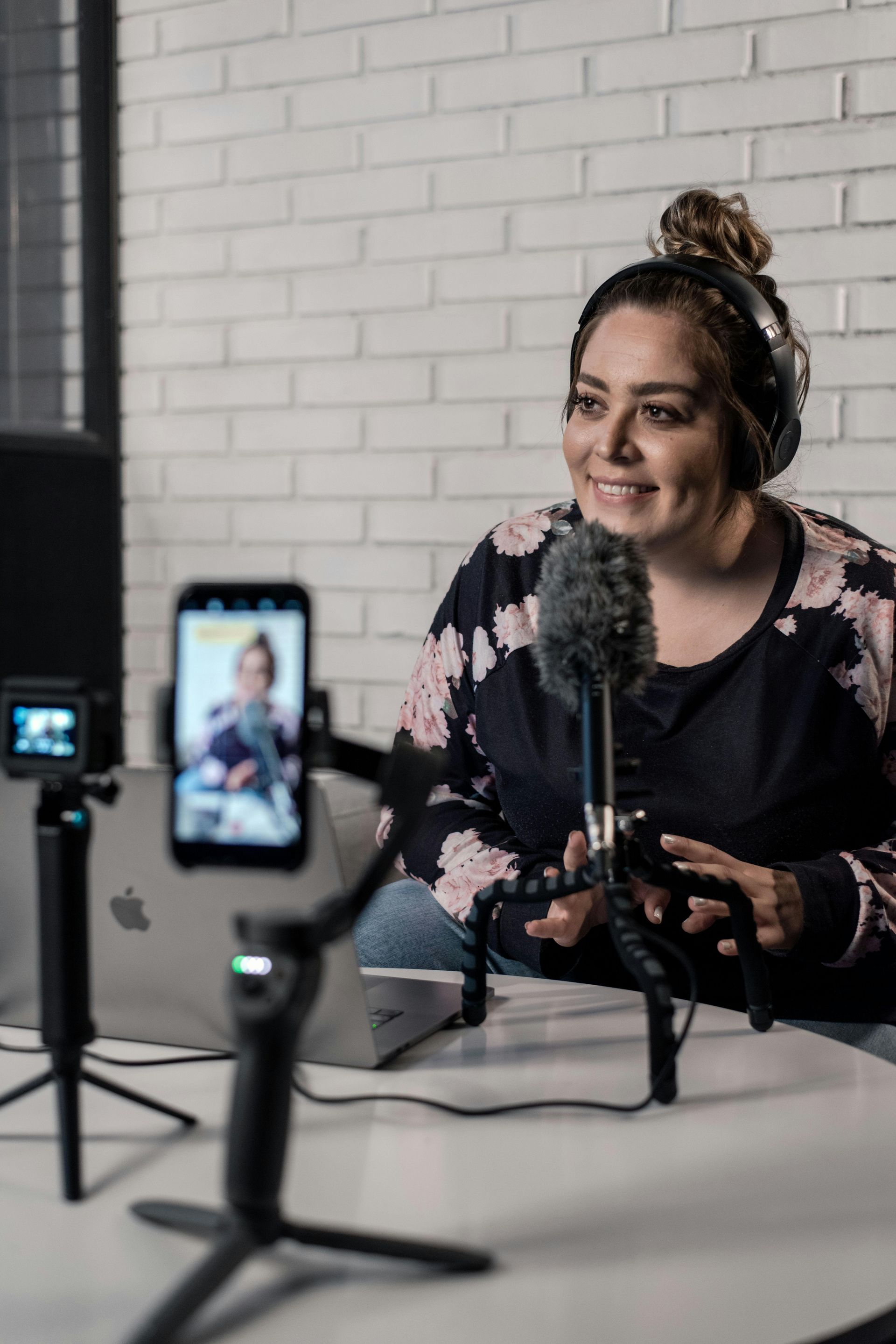 Woman speaking into a microphone during a recorded interview, with a smartphone filming her in a studio.