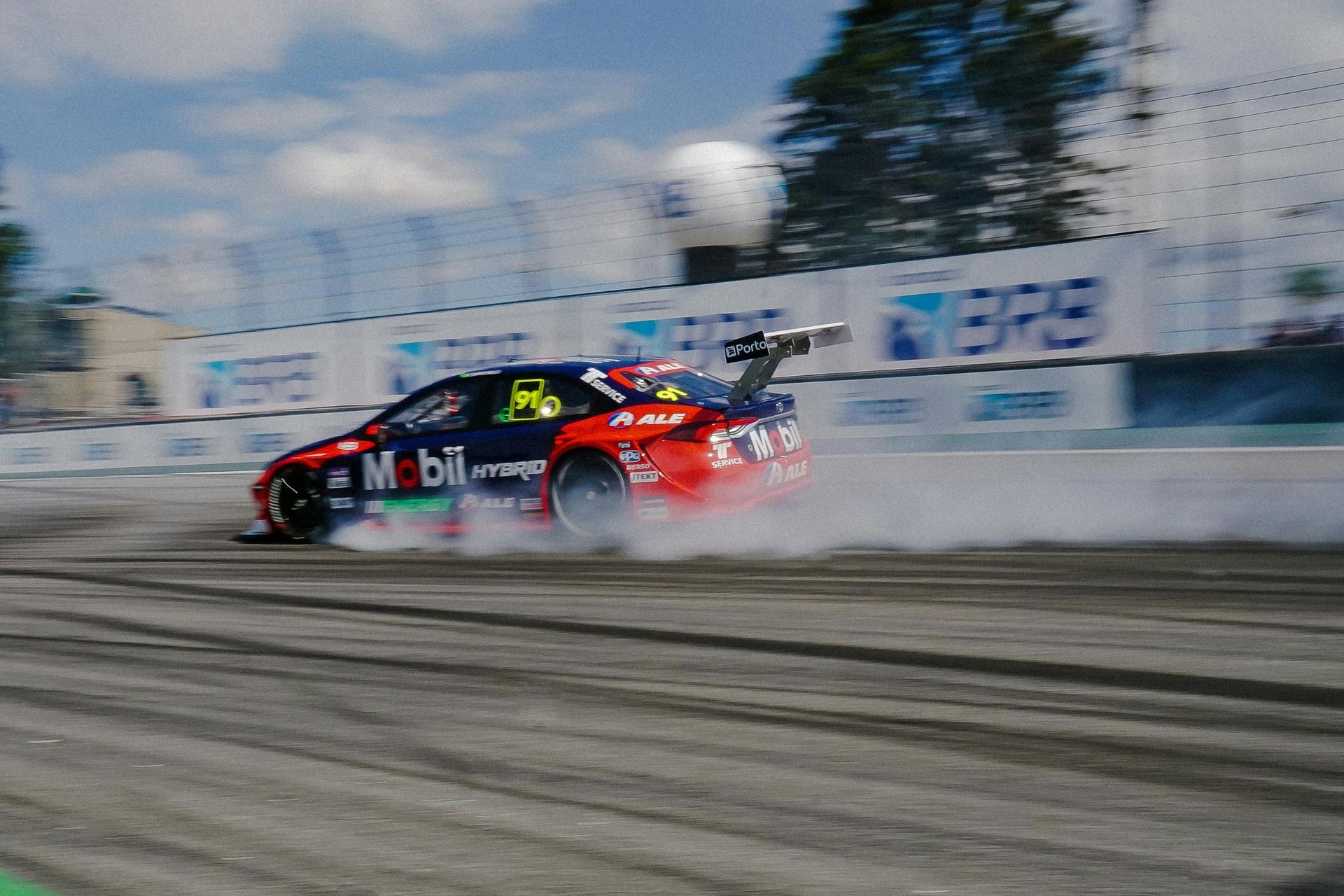 Red race car drifting through smoke on a track, with blurred stands and sky in the background