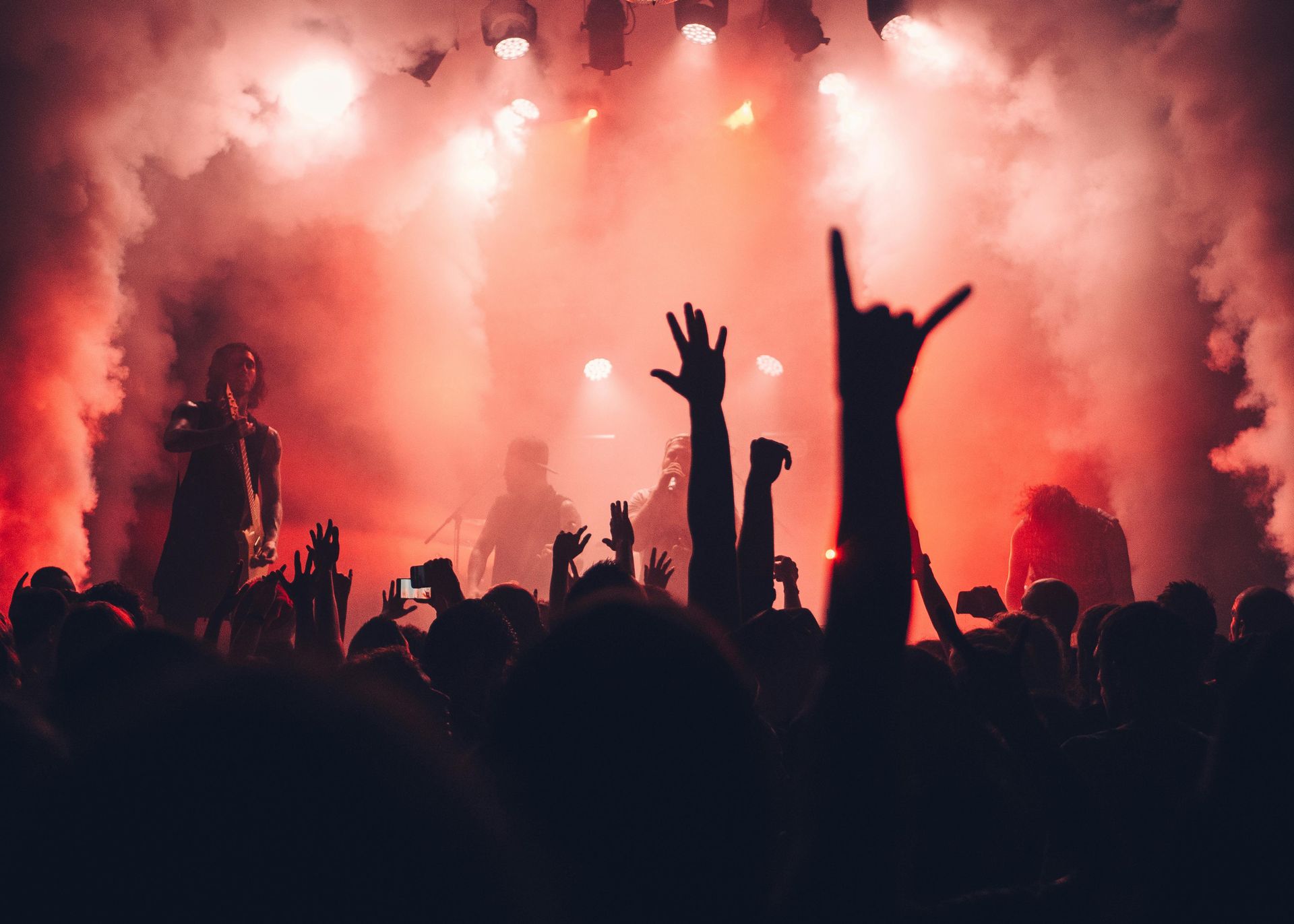 Crowd at a concert with raised hands silhouetted against bright red stage lights and smoke
