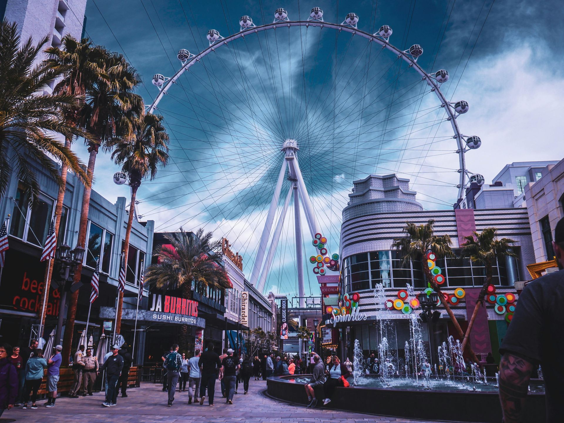 Busy amusement street with a giant Ferris wheel at dusk, framed by palm trees and colorful buildings