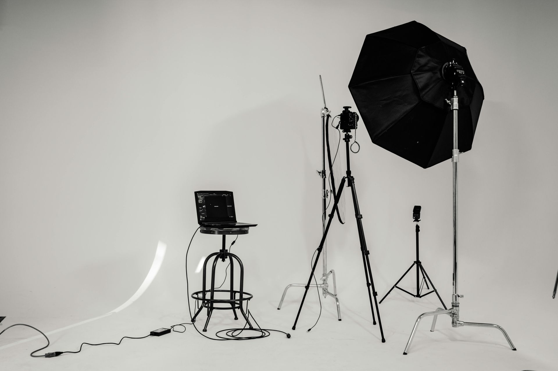 Minimal photography studio with light stands, umbrella softbox, stool, laptop, and cables against a white wall