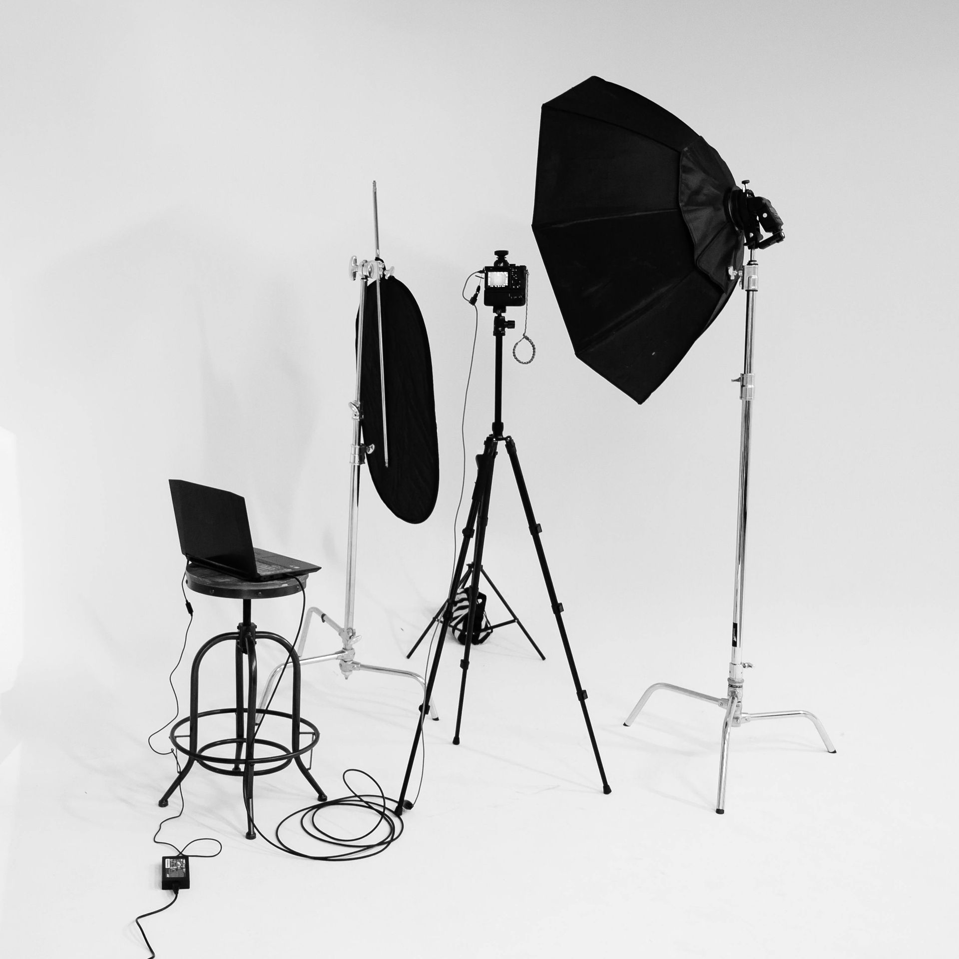 Black-and-white studio setup with tripod camera, light stands, softbox, laptop on stool, and cables