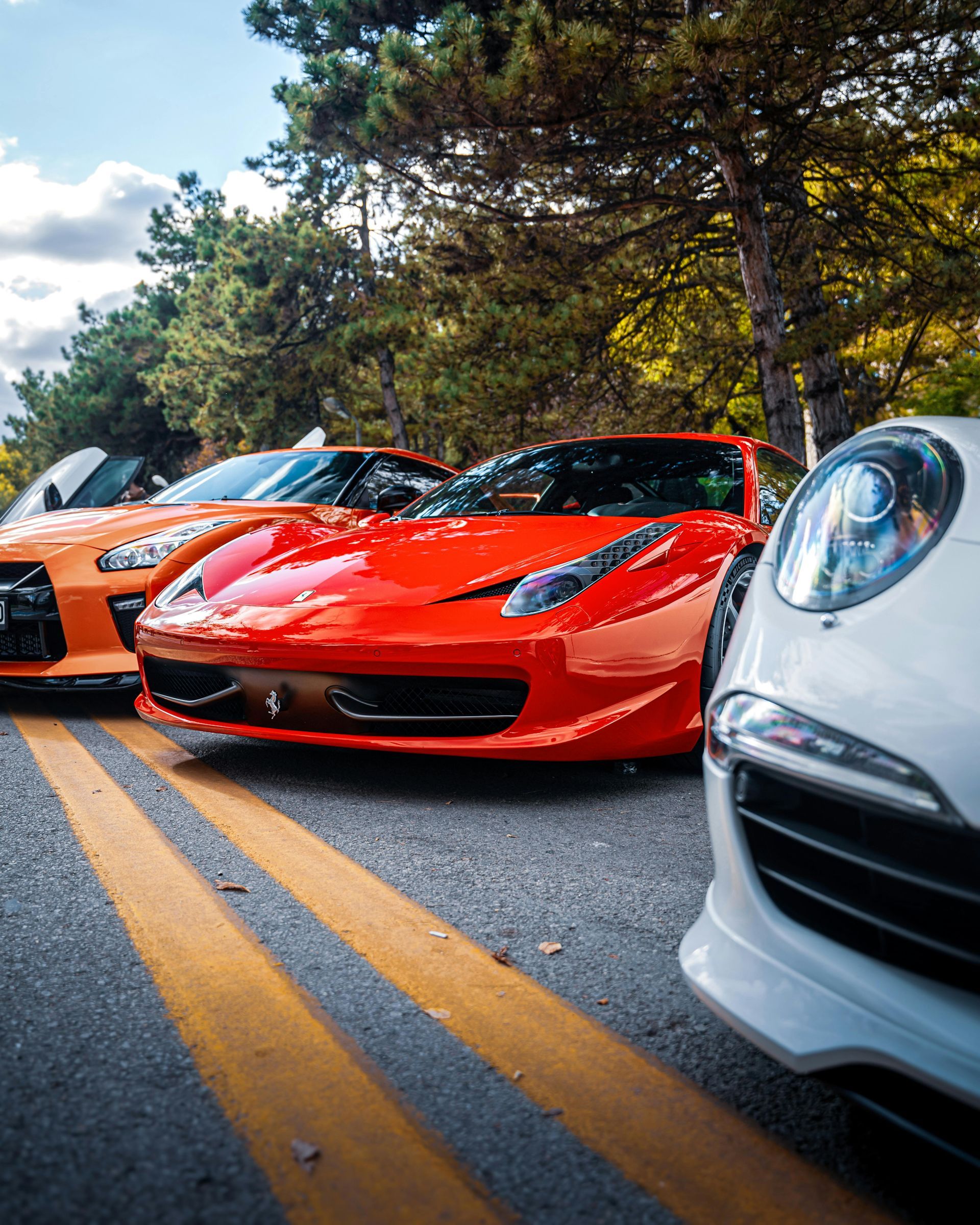 Three sports cars parked on a road, including orange, red, and white cars, viewed from a low angle.