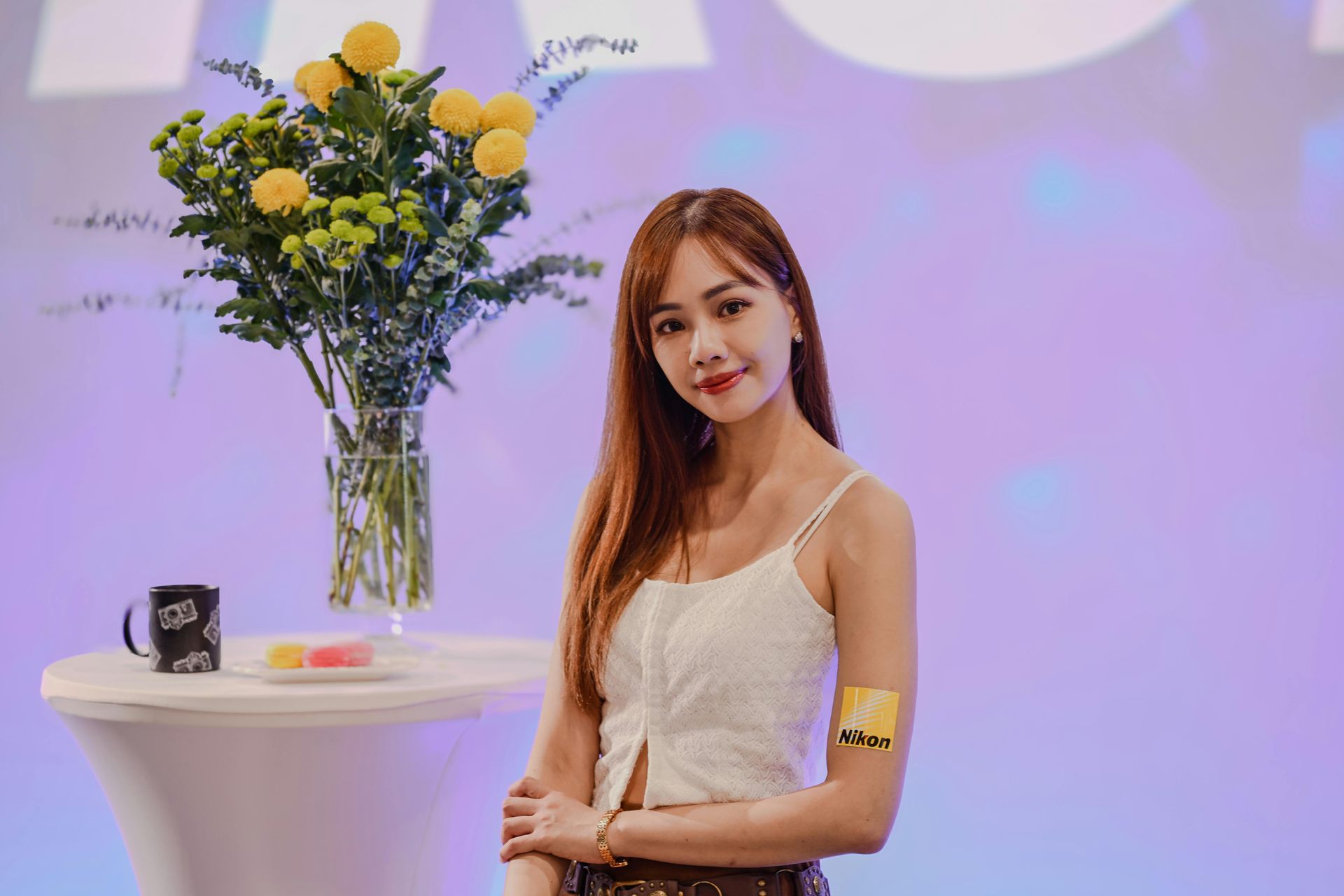 Woman standing beside a table with yellow flowers and a mug in a softly lit event setting