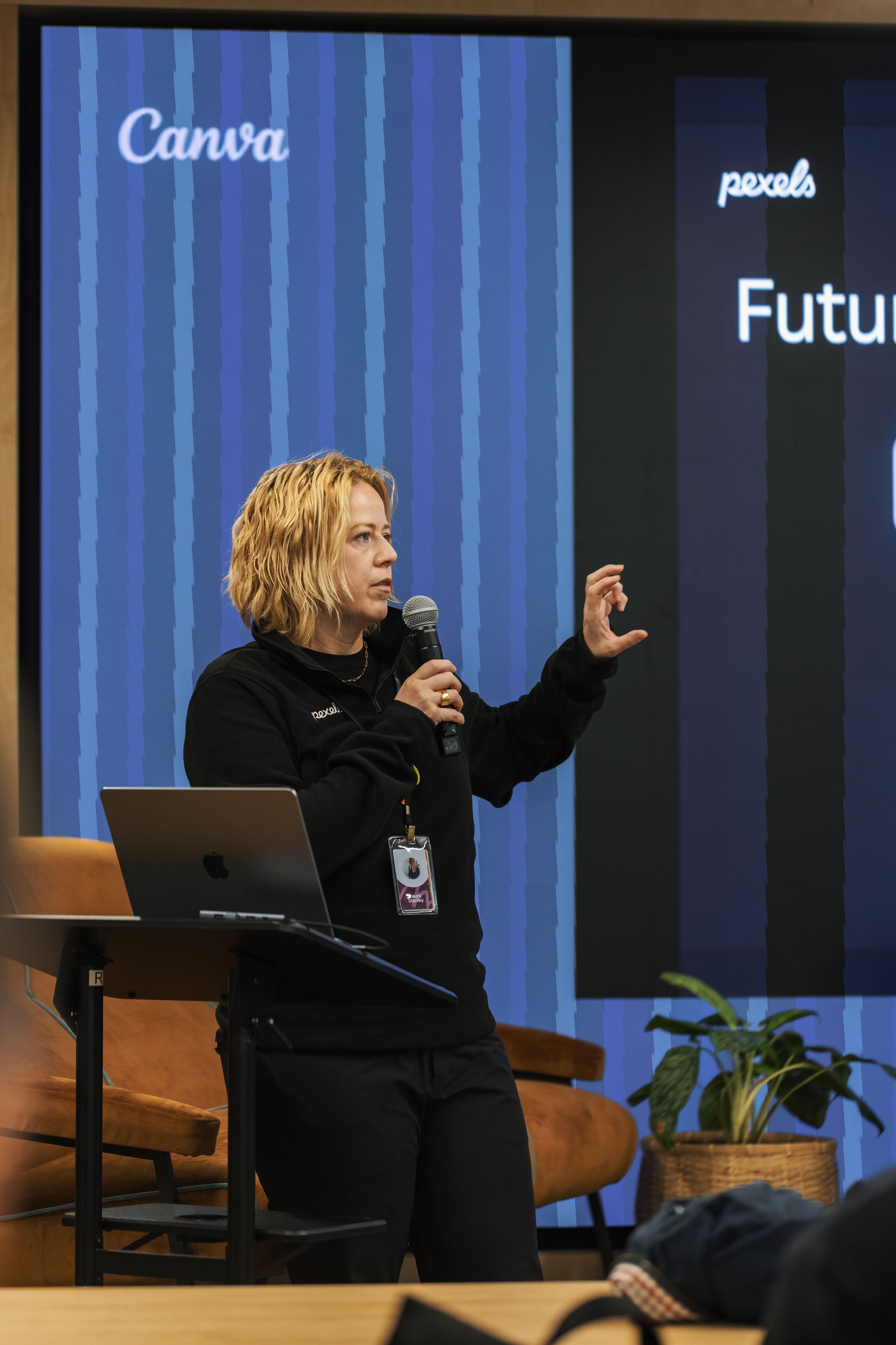 Speaker presenting on stage beside a laptop, gesturing in front of a Canva-branded screen.