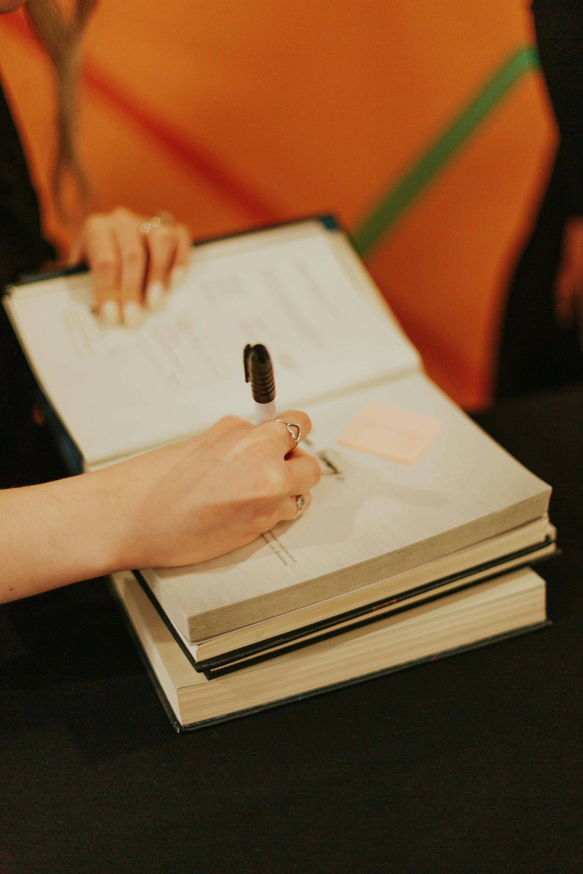 Hand writing in a notebook beside an open book on a dark table